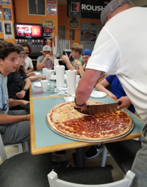 A person slices a large pepperoni pizza at a table with several young people enjoying their meal.