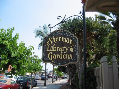 Sign for Sherman Library & Gardens, surrounded by palm trees and greenery, with cars parked along the street.