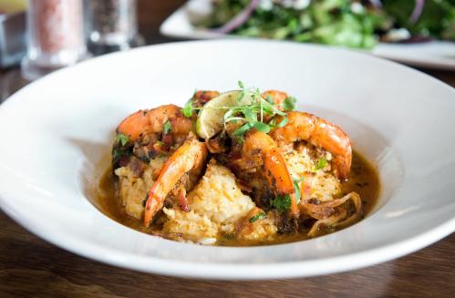 A plate of shrimp served over creamy grits, garnished with lime and herbs, with a fresh salad in the background.