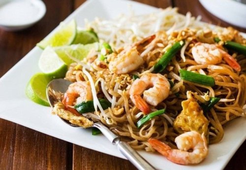 A plate of shrimp stir-fried noodles garnished with lime wedges and bean sprouts, served with a spoon.