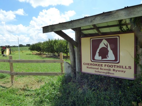 Sign for Cherokee Foothills National Scenic Byway near a field with trees and a blue sky in the background.