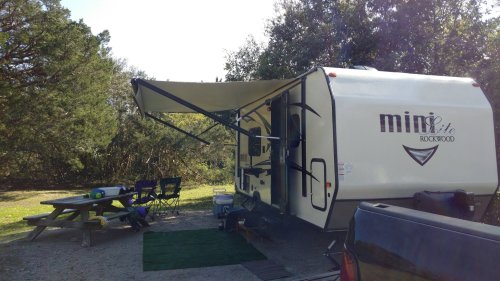 A parked camper with an awning extended, surrounded by trees and a picnic table nearby.