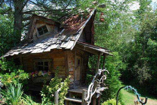 A whimsical, rustic wooden house with a slanted roof, surrounded by greenery and flowers.