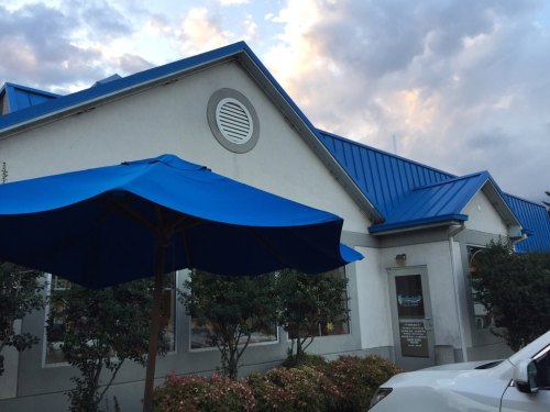 A building with a blue metal roof, featuring a blue umbrella and surrounded by greenery under a cloudy sky.