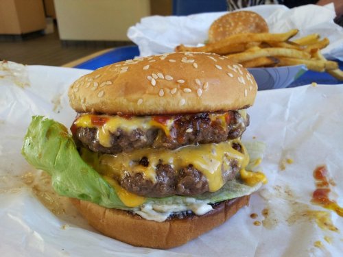 A double cheeseburger with melted cheese, lettuce, and condiments on a sesame seed bun, served with fries in the background.