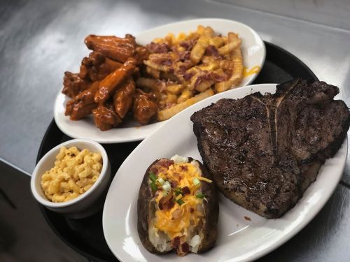 A platter featuring a large steak, loaded baked potato, macaroni and cheese, and wings with fries topped with cheese and bacon.