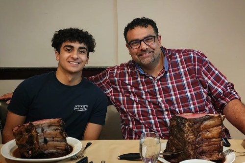 A young man and an older man smile together at a table with large cuts of meat in front of them.