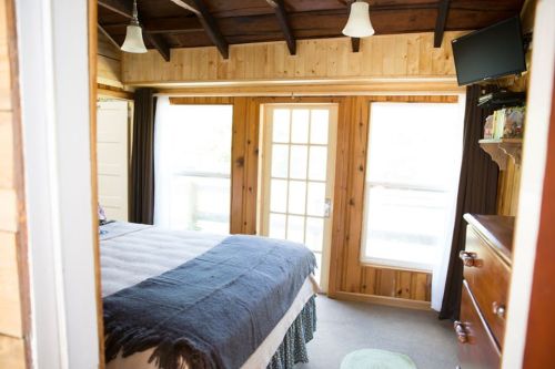 Cozy bedroom with wooden walls, a bed, and large windows letting in natural light. A TV is mounted on the wall.