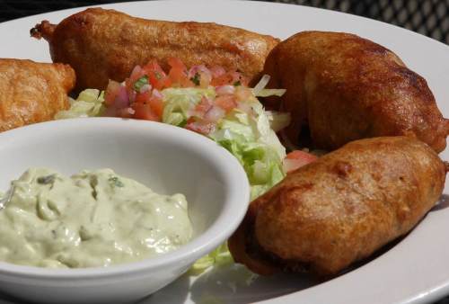 A plate of golden-brown fried snacks, accompanied by fresh lettuce, diced tomatoes, and a bowl of green dipping sauce.