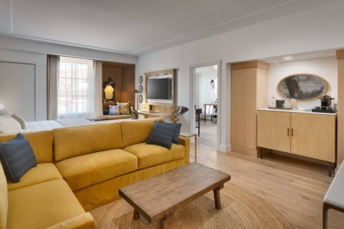 Cozy living area with a yellow sofa, wooden coffee table, and kitchenette, featuring modern decor and natural light.
