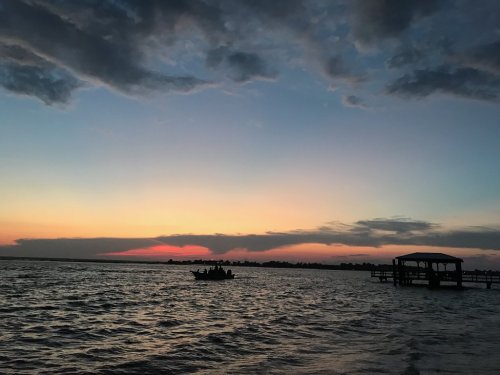 A serene sunset over water, with silhouettes of a boat and a dock against a colorful sky filled with clouds.