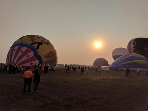 Hot air balloons in a field at sunset, with a crowd of people watching and a hazy sky in the background.