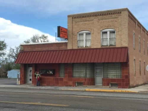 A brick building with a red awning and sign reading "Sid's" on a street, featuring large windows and a small entrance.