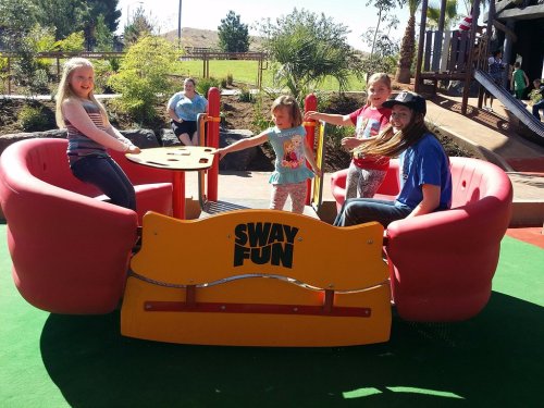 Four children play on a colorful playground device labeled "Sway Fun," surrounded by greenery and other kids in the background.