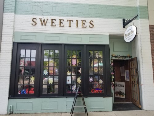 Exterior of a shop named "Sweeties," featuring large windows, floral decorations, and an open sign.