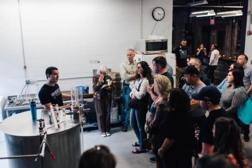 A group of people attentively listens to a speaker in a brewing facility, with equipment and a clock in the background.