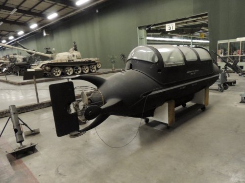 A black submersible displayed in a museum, with military tanks visible in the background.