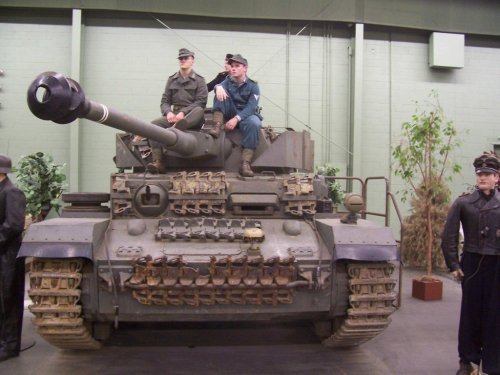 A military tank displayed indoors, with two soldiers in uniform sitting on top and standing beside it.