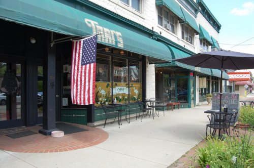 A café with an American flag, outdoor seating, and a sign reading "Sit in the Sunshine" on a sunny day.