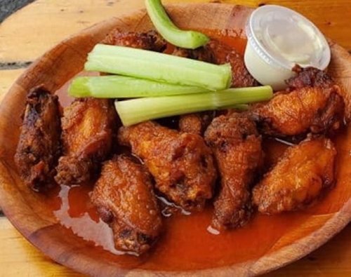 A wooden plate with spicy chicken wings, celery sticks, and a small cup of dipping sauce.