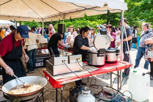 A busy outdoor food festival with vendors cooking and serving food under a large tent, surrounded by a crowd.