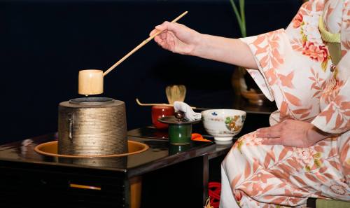 A person in a floral kimono uses a bamboo whisk to prepare tea in a traditional Japanese tea ceremony setting.