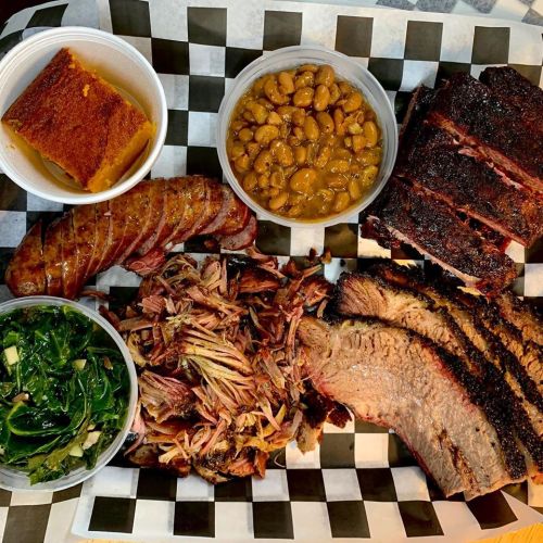 A platter of barbecue featuring brisket, pulled pork, sausage, baked beans, collard greens, and cornbread.