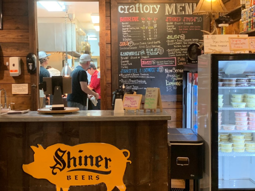 A rustic restaurant interior with a menu board, staff in the kitchen, and a Shiner Beers logo on the counter.