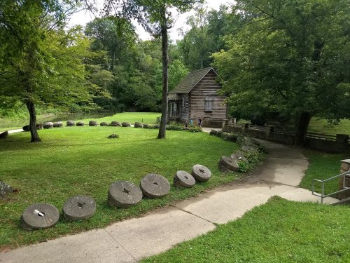 A rustic log cabin surrounded by trees and a grassy area, with stone mill wheels lining a path.