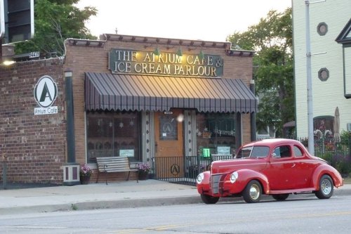 A vintage red car parked in front of The Atrium Cafe & Ice Cream Parlour, a charming brick building at sunset.