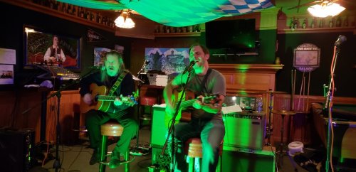 Two musicians perform with acoustic guitars in a lively bar, illuminated by green lights and a festive atmosphere.
