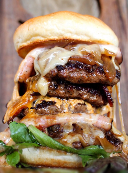 A towering burger with three beef patties, melted cheese, grilled onions, and greens, all sandwiched in a soft bun.