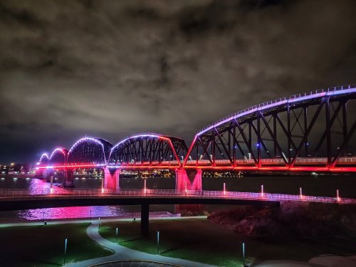 A brightly lit bridge arches over a river at night, illuminated in red and purple against a cloudy sky.
