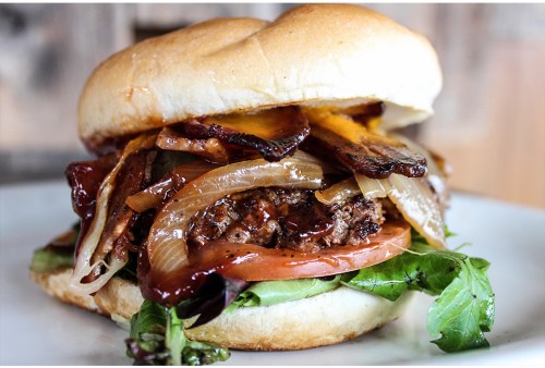 A close-up of a juicy burger with grilled onions, bacon, cheese, lettuce, and tomato on a soft bun.