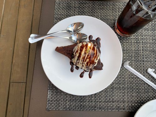 A slice of chocolate cake topped with a scoop of vanilla ice cream and chocolate drizzle, served with two spoons.