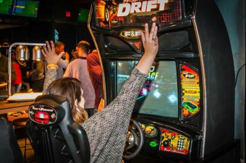 A person celebrates while playing a racing arcade game, hands raised in excitement, with others in the background.