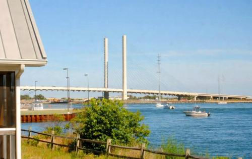 A view of a bridge with tall towers over a calm waterway, framed by greenery and a building in the foreground.