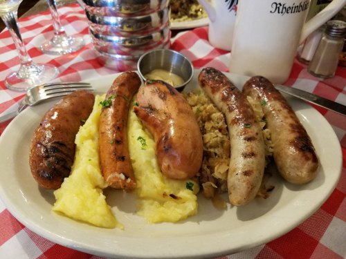 A plate of assorted grilled sausages served with mashed potatoes and sauerkraut, on a checkered tablecloth.