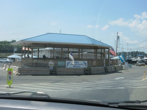 A waterfront restaurant with a blue roof, outdoor seating, and flags, surrounded by boats and a sunny sky.