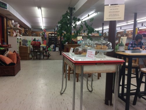 A cozy shop interior with tables, baked goods under glass domes, and various items for sale in the background.