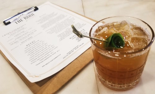 A cocktail with ice and a garnish sits next to a menu on a clipboard, on a marble surface.