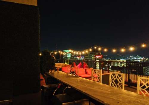 Rooftop lounge at night with string lights, seating, and a city skyline view in the background.