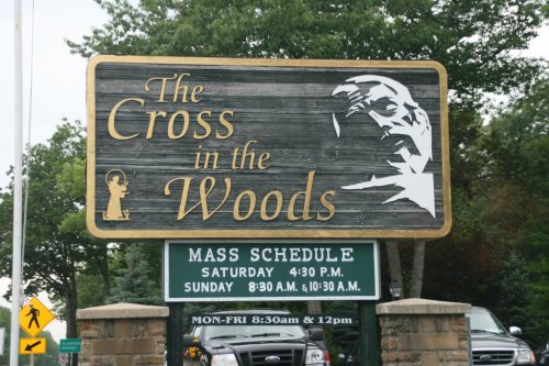 Sign for "The Cross in the Woods" with mass schedule and a silhouette of a religious figure.