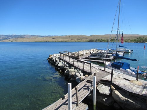 A wooden dock extends into a calm lake, with boats moored and mountains in the background under a clear blue sky.