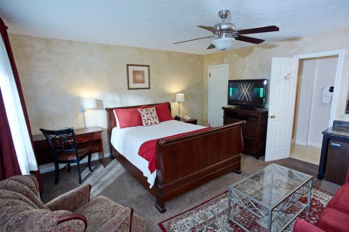 Cozy hotel room featuring a queen bed, seating area, TV, and warm decor with natural light.