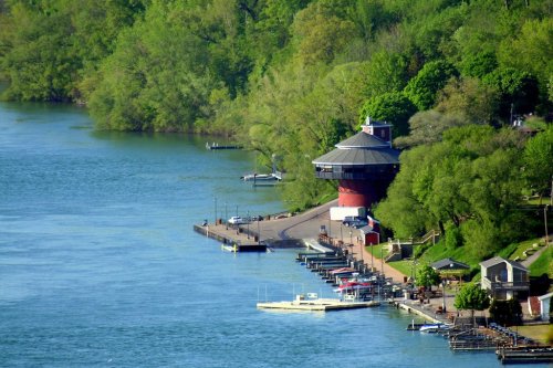 A scenic view of a river with green trees, a round building, and docks lined with boats along the shore.