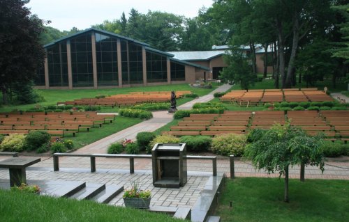 A serene outdoor venue with wooden benches arranged in rows, surrounded by greenery and a modern building in the background.