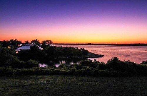 A serene sunset over a calm river, with silhouettes of trees and a building along the shore.