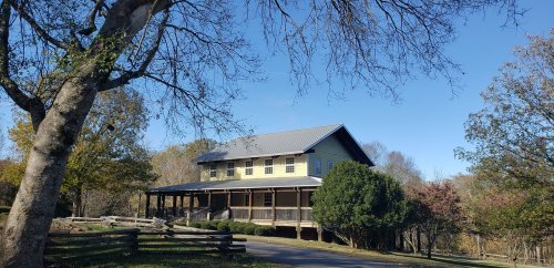 A two-story house with a porch, surrounded by trees and a clear blue sky, set in a rural landscape.