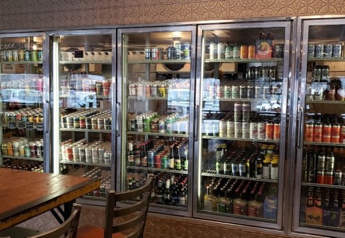 A display of various canned beverages in glass refrigerators, with a wooden table and chairs in the foreground.